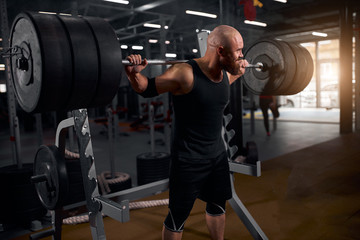 Fototapeta na wymiar Portrait of hairless young bodybuilder training in modern gym, practicing hard, looking away, turning head back, trying to do barbell squats, sportive people concept, close view, portrait