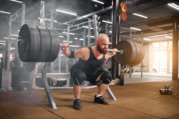 Acctive strong weightlifter sitting with heavy barbell in hands, squatting in modern gym room,...
