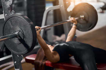 Close up of black iron barbell with heavy discs in hands of strong female weightlifter lying on red bench, attempting to do bench press in modern fitness club, side shot - obrazy, fototapety, plakaty