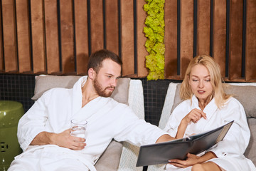Pleasant young couple deciding what drink to order, holding big black menu, looking down, man pointing at menu ready to make order, drinking water, having free time in modern wellness center