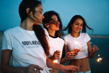 Young happy girls stands on beach with sparkler in sunset light. Teenage girls with sparklers celebrate and laugh. Party, holidays, nightlife and people concept.