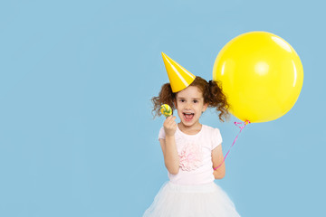 Little pretty curly girl in fashion skirt hold big yellow balloon and lollipop for kids birthday, show excited emotion, over blue background. Space for text.