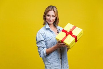 Portrait of happy pretty girl with fair hair in denim casual shirt holding wrapped gift box and looking at camera with smile, enjoying holiday present. indoor studio shot isolated on yellow background