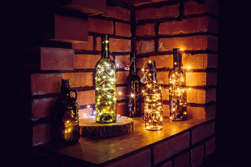Lot of different vintage style beer bottles and green wine bottles decorated with wire string micro led lights on shelf, red brick wall background. Illuminated at night.