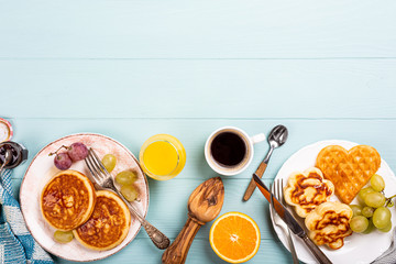 Healthy breakfast background with fresh hot waffles hearts, pancakes flowers with berry jam and fruits on turquoise background, top view, flat lay, copy space.