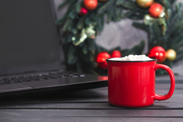 coffee or hot chocolate and marshmallows, next to a spruce Christmas wreath with red and gold balls and a laptop on a gray
