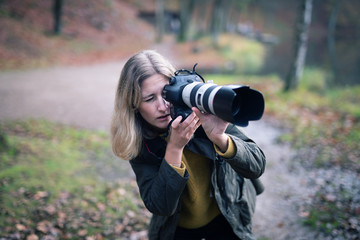 Blonde photographer squatting and using camera with large telephoto lens while taking shots in nature