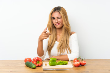 Young blonde woman with vegetables in a table points finger at you