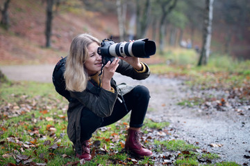 Blonde photographer squatting and using camera with large telephoto lens while taking shots in...