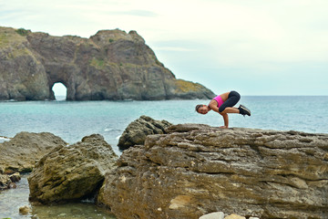 A young athletic woman is doing fitness on a rocky seashore next to a grotto