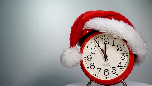 Christmas time concept. Santa Claus hat and big red clock ticking showing twelve hours, on grey background. Waiting for celebration. Time lapse