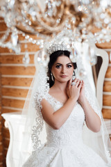 Fototapeta premium Close up portrait of beautiful bride standing by the window at home and tying on earrings. Charming bride in white robe. Beautiful bride with makeup and hair style with crown. 