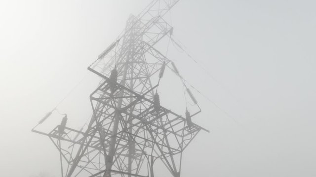 Electricity pylon on misty, foggy morning, tilt down to agricultural farmland hay bales in row across countryside landscape.