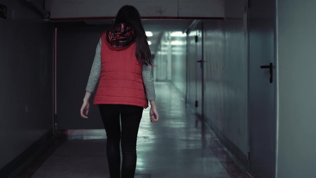 Camera follows young brunette woman in red sleeveless jacket walks quickly along long corridor with low ceilings in the dark.