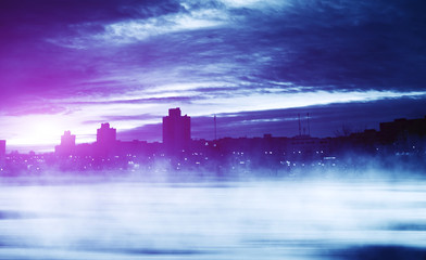 Fototapeta premium Background of winter night city. Snow covered road, fog. Reflections of the neon glow in the sky. Dawn over the winter city