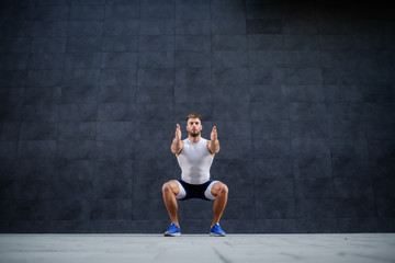 Fototapeta na wymiar Handsome muscular caucasian man in shorts and t-shirt doing squatting exercise outdoors. In background is gray wall.