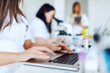 Obraz premium Close up of female lab assistant in white uniform sitting in lab and using laptop for data entry. In background is her colleague working.