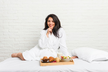 Young curvy woman taking a breakfast on the bed smiling happy and confident, touching chin with...