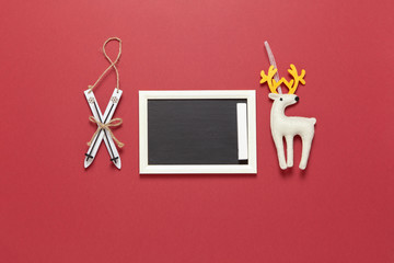 Christmas toys wooden skis, white felt toy deer and small clean chalk board with copy space on dark red background. Festive, New Year concept. Horizontal, flat lay. Minimal style. Top view