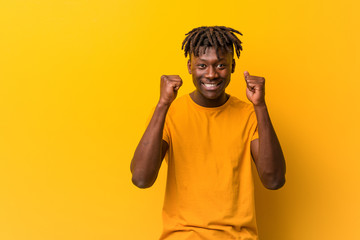 Young black man wearing rastas over yellow background raising fist, feeling happy and successful....