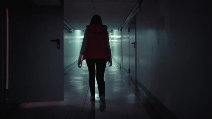 Back view young brunette woman in a red sleeveless jacket walks quickly along a long corridor with low ceilings in the dark, tracking shot. - Powered by Adobe