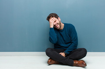 young handsome man sitting on floor laughing and slapping forehead like saying d‚Äôoh! I forgot...
