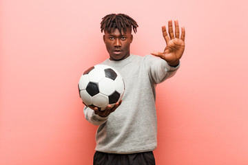 Young fitness black man holding a soccer ball standing with outstretched hand showing stop sign,...