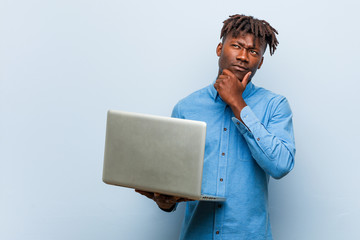 Young rasta black man holding a laptop looking sideways with doubtful and skeptical expression.