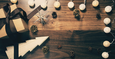 Present paper box wrapped with brown ribbon on wooden background with garland, decorative christmas tree and pine cones. Celebration and holiday concept.