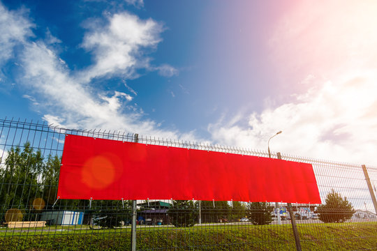 Red blank advertising banner hanging on a fence against the blue sky on a sunny day.