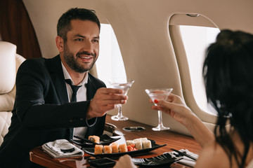 cropped view of woman holding martini glasses near happy man in private jet