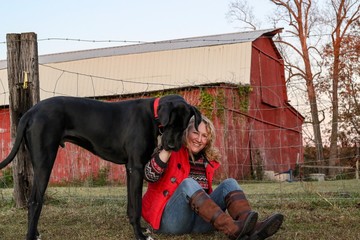 Portrait of a middle aged active country woman and her black Great Dane dog sitting on a grassy field with a red barn in background 