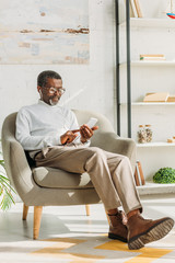 stylish african american man sitting in armchair, listening music in erphones and using digital tablet