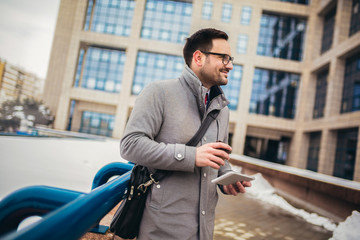 Confident young man in glasses drinking coffee outdoors and using digital tablet