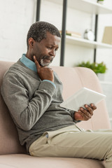 thoughtful african american man sitting on sofa and using digital tablet