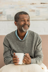 smiling african ameriican man sitting with blanket on knees and holding cup of warming drink