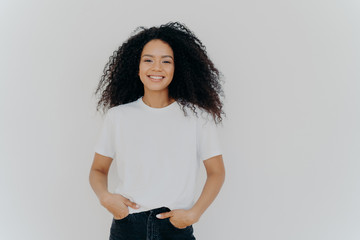 Isolated shot of young African American woman wears white t shirt, expresses good emotions, stands...