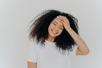 Photo of happy lady keeps hand on forehead, tilts head, smiles joyfully, keeps eyes closed, has curly Afro hair, wears casual white t shirt, enjoys pleasant moment in life, feels little bit shy