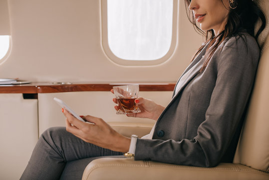 cropped view of businesswoman in formal wear holding glass with whiskey and smartphone in private jet - Powered by Adobe