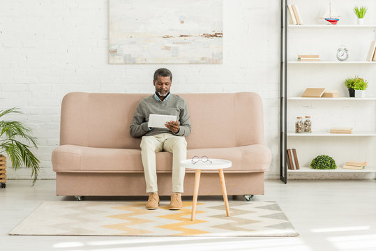 senior african american man sitting on sofa in living room and using digital tablet