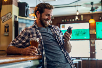 Smiling man with beer glass using smartphone at bar counter