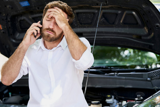 Frustrated man talking on mobile phone while leaning against broken car