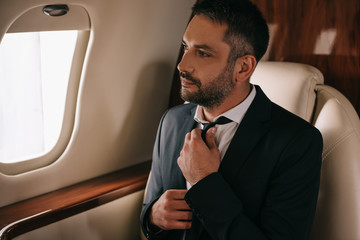 bearded businessman touching tie and sitting in plane