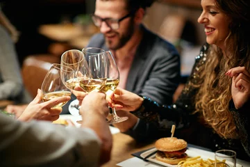 Foto op Canvas Beste vrienden die in restaurant zitten voor het diner en een toast maken met witte wijn. Op tafel staat eten. © chika_milan