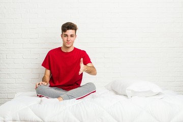 Young teenager student man on the bed stretching hand at camera in greeting gesture.