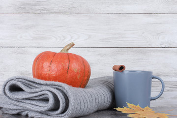 Grey coffee cup, cinnamon, yellow leaves, stack of woolen plaid and pumpkin over white wooden wall background. Autumn drink concept. Fall, coffee, hot drink, thanksgiving, side view, copy space