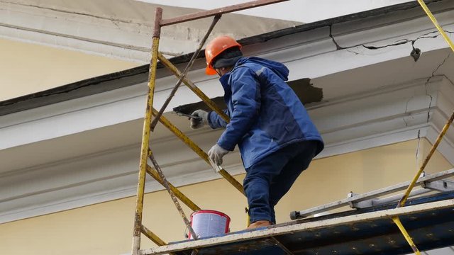 A construction worker on a scaffolding plaster decor on a building. Daytime. Bucket with putty. Protective orange helmet on the head