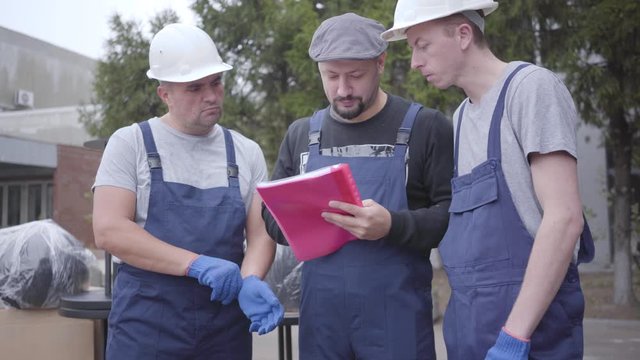 Two adult Caucasian movers in helmets and blue uniform standing with their chief. Three workers reading waybill or consignment, planning work.
