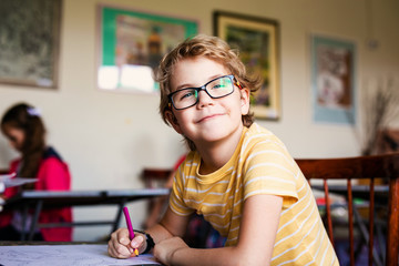 Blonde boy with glasses drawing. Group of elementary school pupils in classroom on art class....