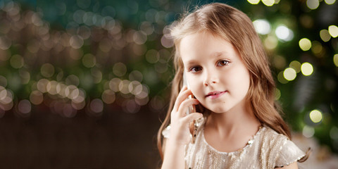 Portrait of a smiling long-haired little girl in dress on background of  lights. Little girl talking on the phone. Christmas, New Year and birthday celebration concept. Winter holidays. Copy space